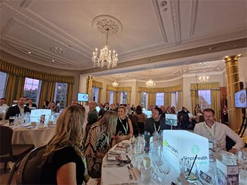People seated at elegantly decorated tables in a formal dining room with chandeliers, engaged in conversation during an event.