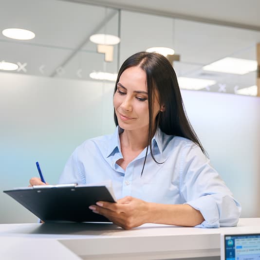 A woman in a light blue shirt writes on a clipboard at a desk in a bright, modern room.