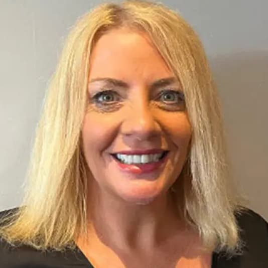 Portrait of a smiling woman with shoulder-length blonde hair, wearing a black top, against a plain background.