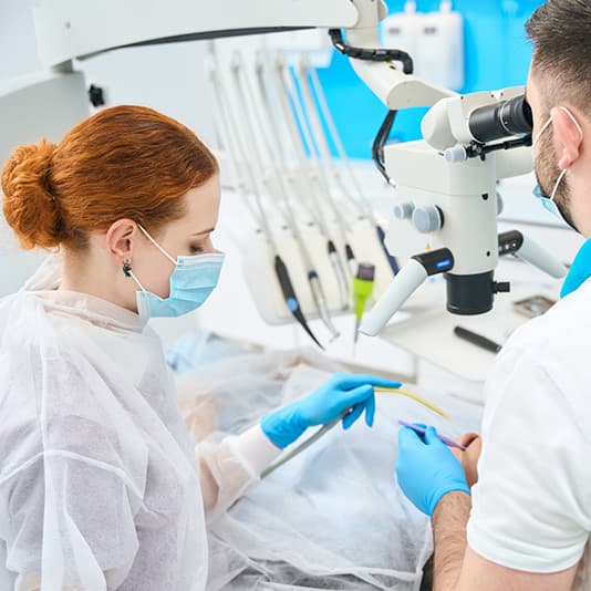 Two dental professionals in masks and gloves use a microscope and tools during a dental procedure in a clinic.