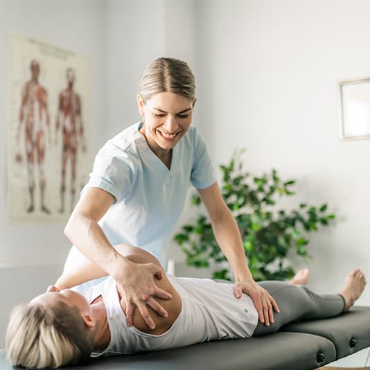 A physical therapist assists a patient with a shoulder stretch on a treatment table in a clinic, with anatomical charts and a plant in the background.