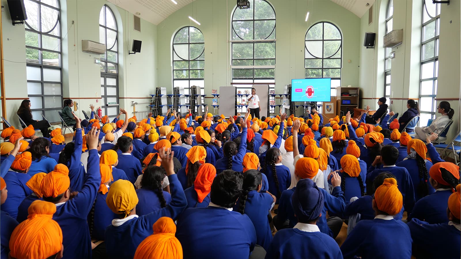 Students in blue uniforms and orange head coverings sit in a hall, raising hands toward a speaker near a screen. Large windows in the background.