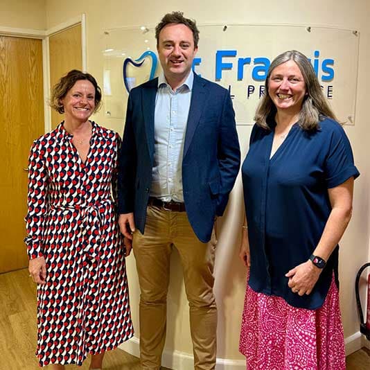Three people smiling, standing in an office in front of a sign. The two women wear patterned dresses, and the man wears a blazer and khakis.