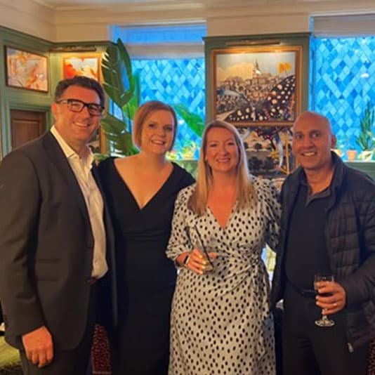 Denplan staff smiling and posing together at an indoor Dine with Denplan event. Two women in dresses and two men in suits. Artwork and plants in the background.