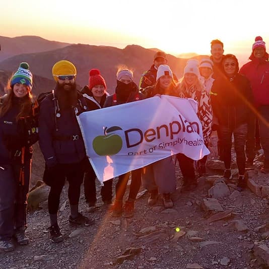 Group of people on a mountain at sunrise, holding a "Denplan" banner, dressed in winter gear, smiling, with distant peaks visible.
