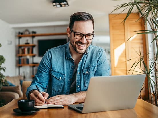 Smiling man in glasses works on a laptop at a wooden table, writing in a notebook. Cozy room with plants and shelves in the background.