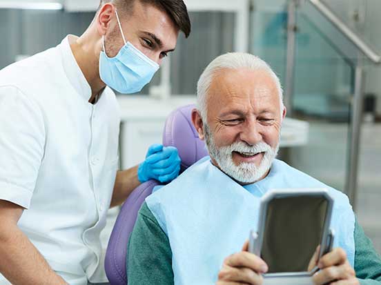 Elderly man sitting in a dental chair, smiling while looking into a handheld mirror, with a dentist beside him wearing a mask and gloves.