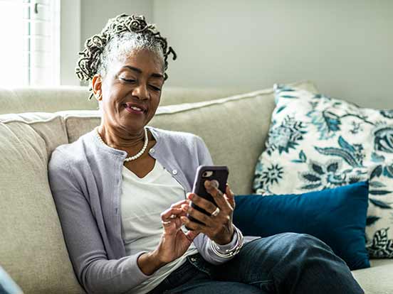 Woman with braided hair, wearing a cardigan, sitting on a sofa, smiling while using a smartphone. Decorative pillows are in the background.