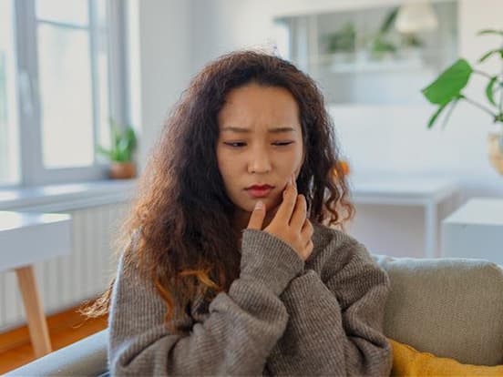 Woman with curly hair in a gray sweater looks in pain whilst holding her face, sitting on a couch in a bright room with plants and large windows.
