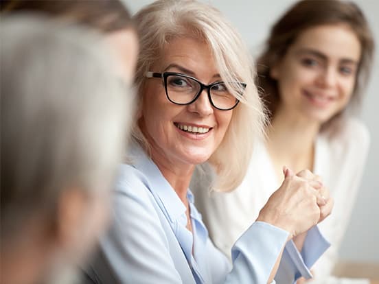 Smiling woman with glasses and white hair sits at a table, engaged in conversation with others, who are slightly out of focus.
