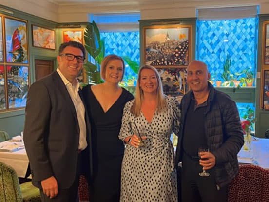 Four people smiling and posing together in a warmly lit restaurant, with colorful artwork and plants in the background.