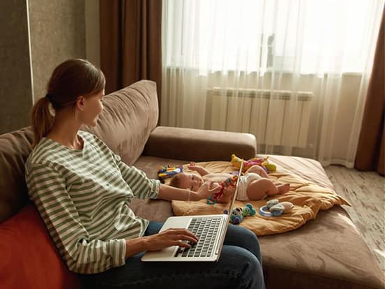 A person on a sofa using a laptop, with a baby lying on cushions nearby, surrounded by toys.