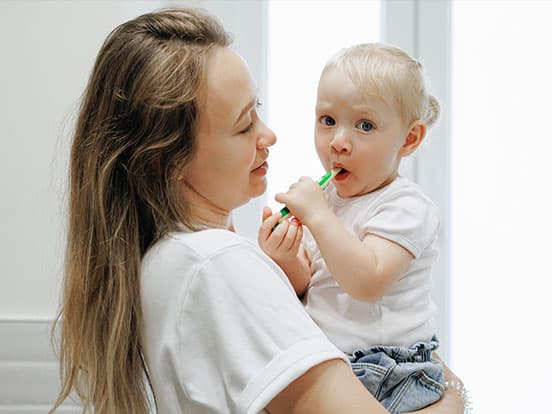 Woman holding a toddler who is brushing their teeth with a green toothbrush, both wearing white shirts in a bright room.